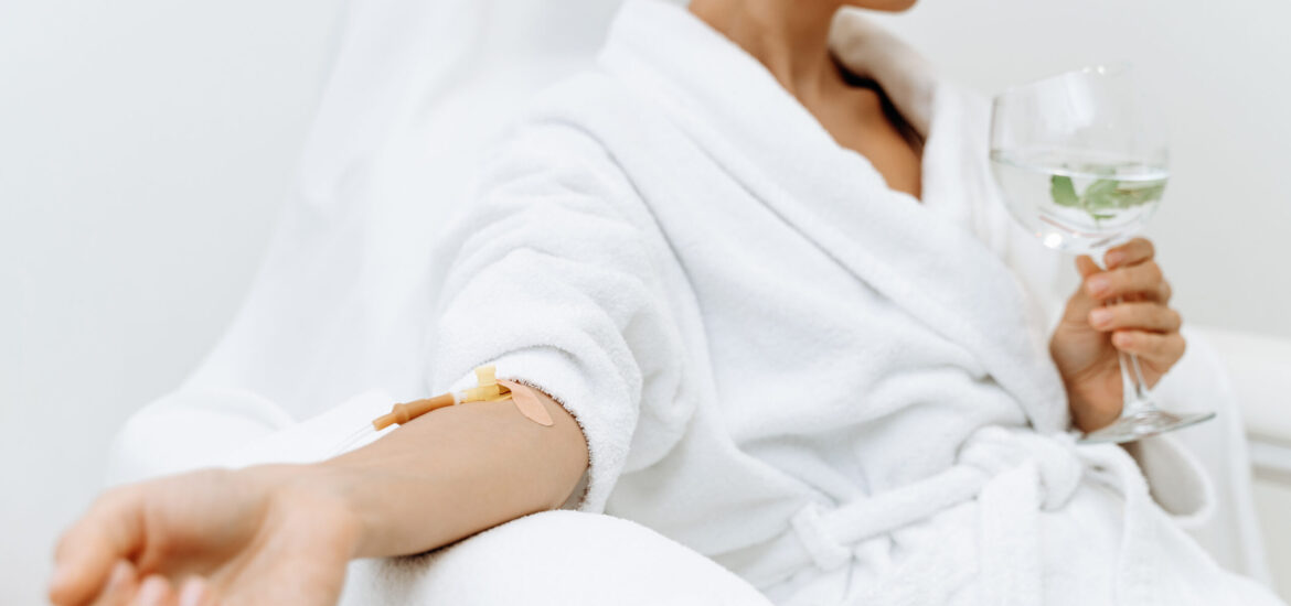 Close up of calm female patient sitting with tube and needle during IV infusion. Girl holding glass with lemon water and relaxing