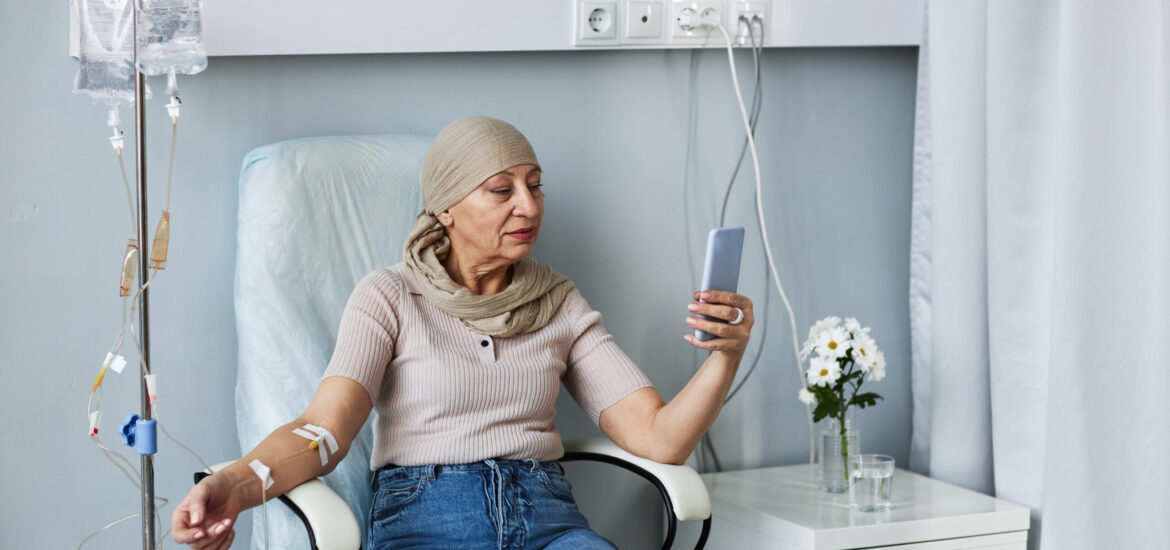 Portrait of senior woman sitting in comfortable chair with IV drip and using smartphone during chemotherapy treatment