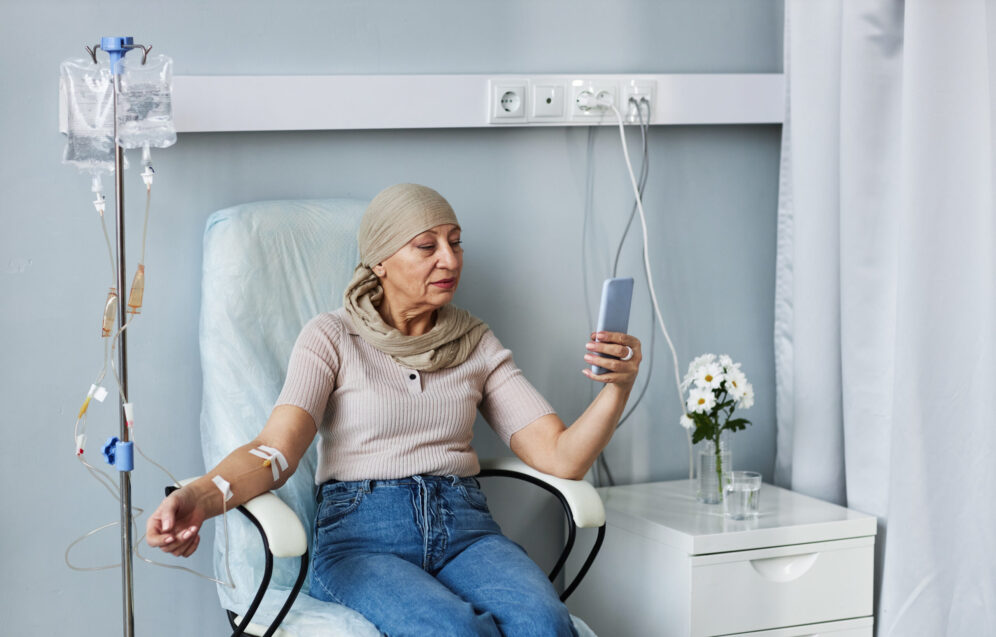 Portrait of senior woman sitting in comfortable chair with IV drip and using smartphone during chemotherapy treatment